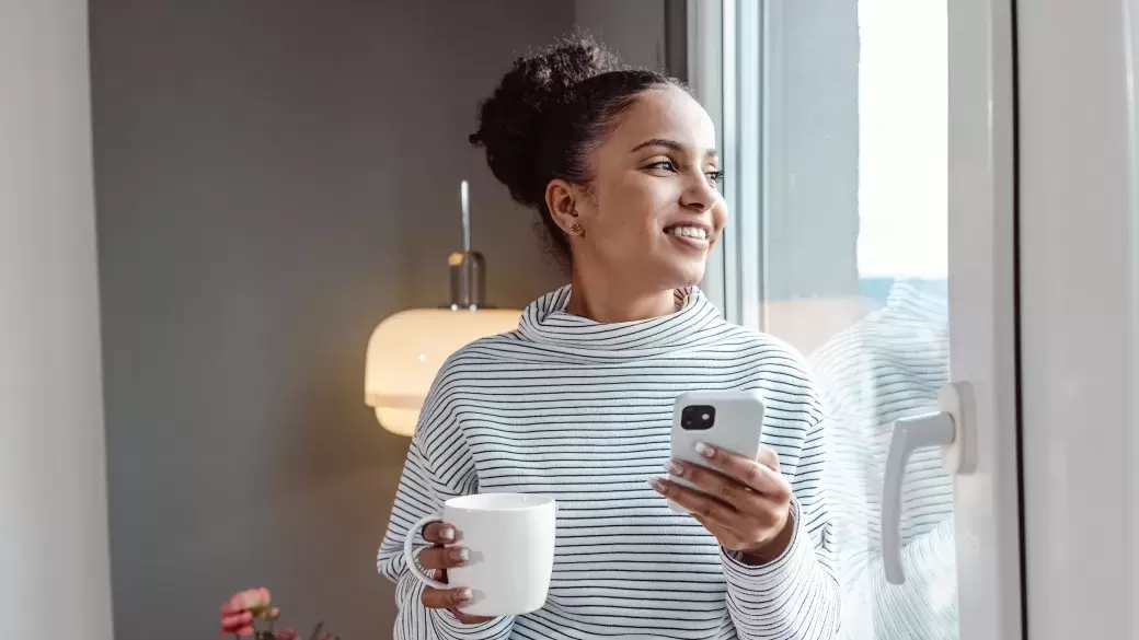 Happy woman standing in window of new phone holding drink and phone