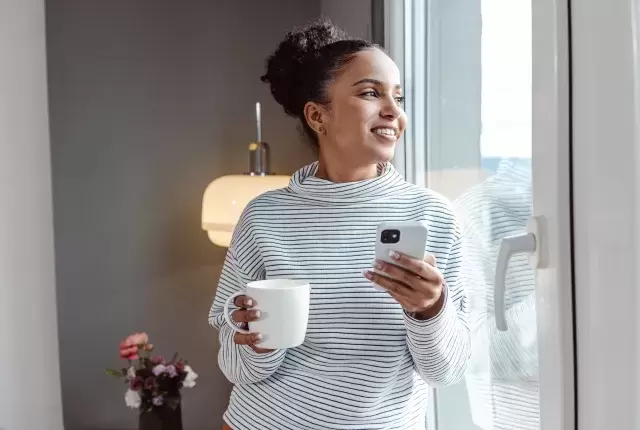 Happy woman standing in window of new phone holding drink and phone